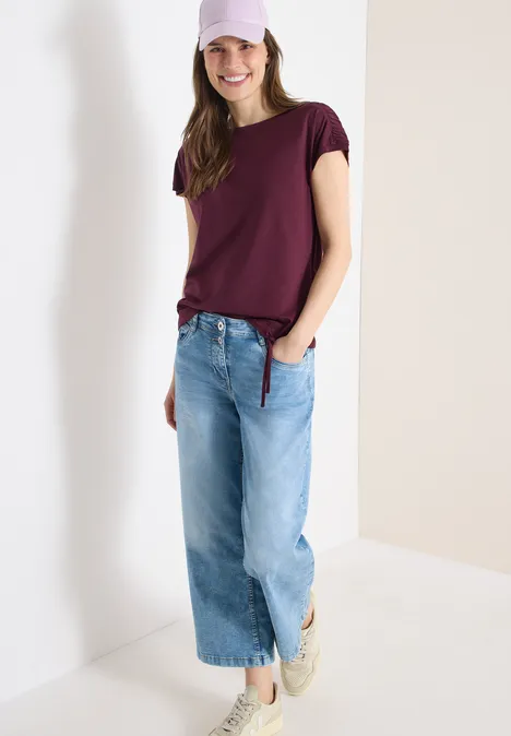 Eine Person in einem dunkelroten T-Shirt, weitem, hellblauen Jeanshose und weißen Sneakern, lässig an eine Wand gelehnt.